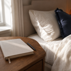 A blank notebook and pen sit on a bedside table beside a neatly made bed in soft morning light.