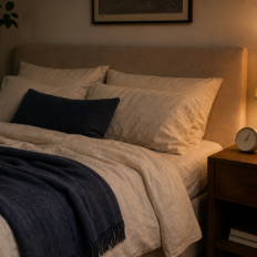 A calm bedroom with cream bedding, a navy throw, a lamp, a face-down phone, and a plain analog alarm clock on the nightstand.