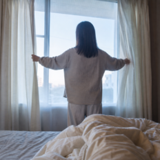 An adult seen from behind opening bedroom curtains to soft natural morning light.
