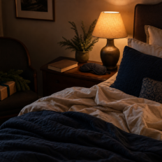 A tidy bedroom with navy and cream bedding, a warm bedside lamp, a sleep mask, a closed book, and a small Christmas gift near a chair.