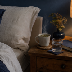 A bedside table with a mug of tea, a glass of water, a closed book, and a warm lamp beside cream bedding in a tidy bedroom.