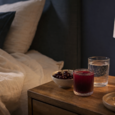 A bedside table holds a red drink, a bowl of cherries, a glass of sparkling water, and an unbranded bottle beside cream bedding.